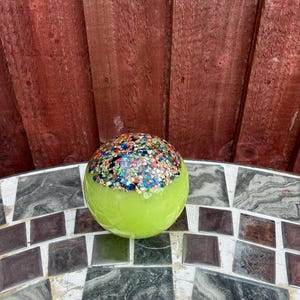 May include: A spherical glass paperweight featuring a smooth, translucent lime green base and a textured top section embedded with a dense cluster of multicoloured, reflective glass shards in shades of blue, red, yellow, and silver.