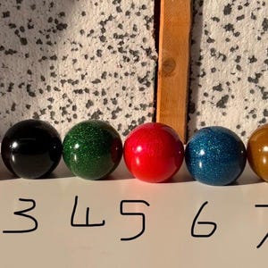 May include: Eight spherical knobs arranged in a row, labelled 1 through 8. The colours are silver, orange, black, green, red, blue, brown, and teal. Several of these knobs feature a glittery finish, while others have a solid or translucent appearance.