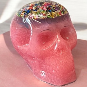 May include: A translucent pink resin skull sculpture featuring a smooth, glossy finish. The top portion of the skull is embedded with a dense cluster of multicoloured metallic foil flakes in shades of blue, gold, green, and red, creating a vibrant contrast.