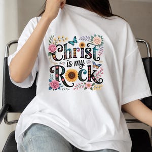 May include: A white short-sleeved t-shirt featuring a colorful graphic print on the chest. The design includes the black text Christ is my Rock, surrounded by illustrations of pink roses, a yellow sunflower, and two blue butterflies.