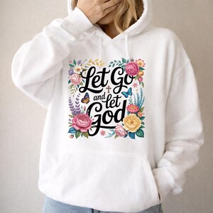 May include: A white hooded sweatshirt featuring a central graphic with the black text Let Go and let God, surrounded by colorful illustrations of pink, yellow, and red flowers, green leaves, a small brown cross, and two blue and orange butterflies.