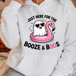May include: A white hooded sweatshirt featuring a graphic of a white ghost wearing heart-shaped sunglasses while floating in a pink flamingo pool float. The text reads JUST HERE FOR THE BOOZE & BOOS, surrounded by small yellow star accents.