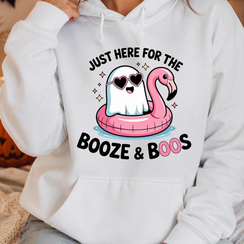 May include: A white hooded sweatshirt featuring a graphic of a white ghost wearing heart-shaped sunglasses while floating in a pink flamingo pool float. The text reads JUST HERE FOR THE BOOZE & BOOS, surrounded by small yellow star accents.
