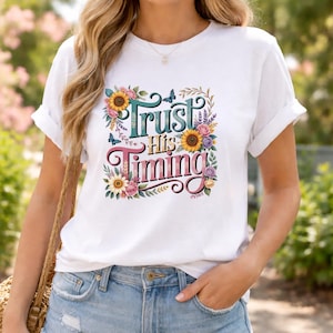 May include: A white short-sleeved t-shirt featuring a colorful graphic print of sunflowers, roses, and small blue butterflies surrounding the text "Trust His Timing" in a decorative font. The design is centered on the chest of the garment.