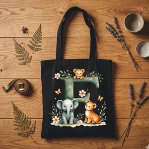 May include: Black tote bag featuring a whimsical design with the letter "E" adorned with an elephant, a lion cub, and floral accents. The bag has two black handles and is made of a durable fabric.
