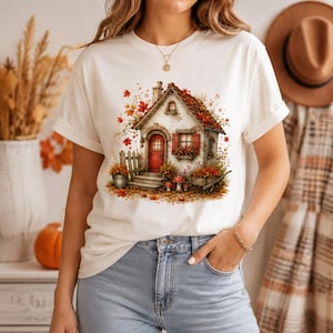 May include: A white short-sleeve t-shirt featuring a graphic print of a cottage with a red door, brown roof, and autumn foliage. The design includes a wooden fence, a wheelbarrow filled with leaves, and small mushrooms on the ground.