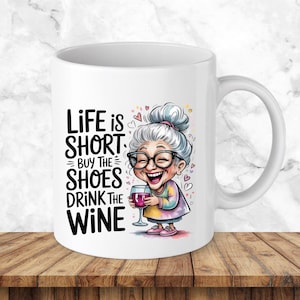 May include: A white ceramic mug featuring a cartoon illustration of an elderly person with grey hair, glasses, and a colourful shirt holding a glass of red wine. The text reads: Life is short, buy the shoes, drink the wine, surrounded by small hearts.