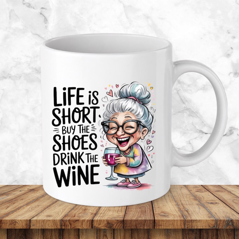 May include: A white ceramic mug featuring a cartoon illustration of an elderly person with grey hair, glasses, and a colourful shirt holding a glass of red wine. The text reads: Life is short, buy the shoes, drink the wine, surrounded by small hearts.