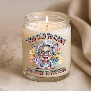 May include: A clear glass jar candle with a lit wick, featuring a graphic of an elderly person with grey hair, glasses, and a purple cardigan sticking their tongue out. The text reads TOO OLD TO CARE TOO TIRED TO PRETEND, surrounded by colourful hearts.