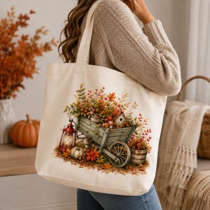 May include: A cream-colored canvas tote bag featuring a printed illustration of a rustic wooden cart overflowing with autumn foliage, pumpkins, a red lantern, and a small birdhouse, all set against a backdrop of vibrant fall colors and leaves.