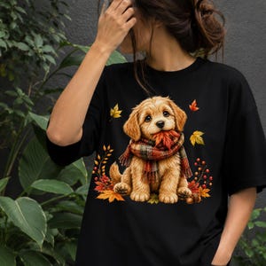 May include: A black t-shirt featuring a graphic print of a golden-brown puppy wearing a plaid scarf and holding an autumn leaf in its mouth, surrounded by colorful fall foliage, red berries, and small acorns on the front of the garment.