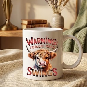 May include: A white ceramic mug featuring a colorful illustration of a Highland cow with long horns and shaggy fur. The text printed on the mug reads: WARNING: FREQUENT MOO-D SWINGS. The mug has a standard handle and a smooth, glossy finish.