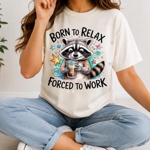 May include: A cream-coloured t-shirt featuring a graphic of a raccoon holding a drink. The text above reads BORN TO RELAX and the text below reads FORCED TO WORK. The design includes colourful stars and swirling accents in blue, pink, and yellow.