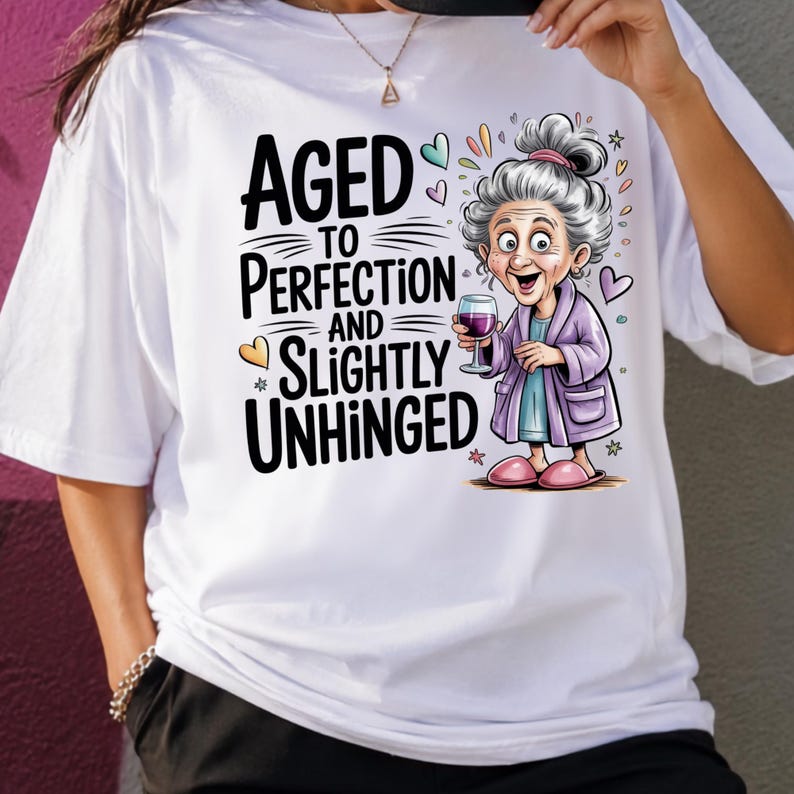 May include: A white t-shirt featuring a graphic of an elderly person with grey hair in a purple robe holding a wine glass. The text reads AGED TO PERFECTION AND SLIGHTLY UNHINGED, accompanied by small colourful heart and star illustrations.
