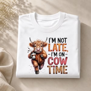 May include: A white t-shirt featuring a graphic of a brown Highland cow holding a coffee cup. The text reads, I'M NOT LATE, I'M ON COW TIME. The cow has shaggy fur and horns, with coffee splashes around the cup. The design is centered on the chest.