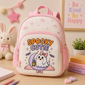 May include: A light pink and cream backpack featuring a cartoon ghost holding a lantern on a crescent moon with the text SPOOKY CUTIE. The bag has a top handle, adjustable straps, side mesh pockets, and a front zippered compartment with bow accents.