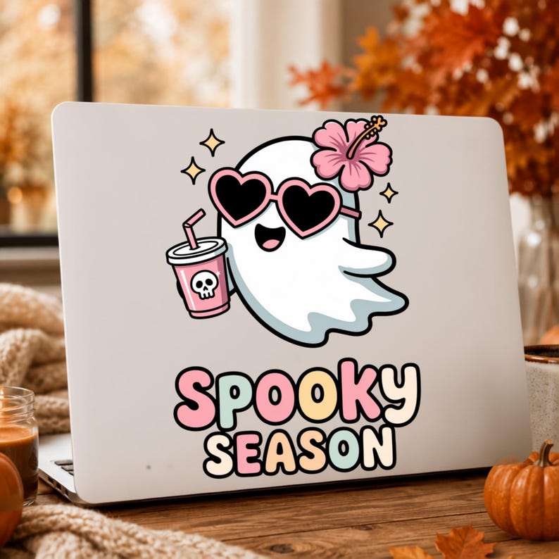 May include: A white laptop decal featuring a cartoon ghost wearing pink heart-shaped sunglasses and a pink hibiscus flower. The ghost holds a pink cup with a skull icon and a straw. Below the ghost, the text reads Spooky Season in pastel letters.