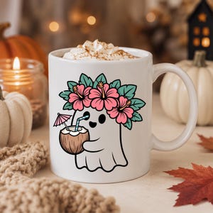 May include: A white ceramic mug featuring a graphic of a ghost wearing a pink hibiscus flower crown and holding a coconut drink with a straw and a small pink umbrella. The mug is filled with whipped cream topped with a dusting of brown spice.