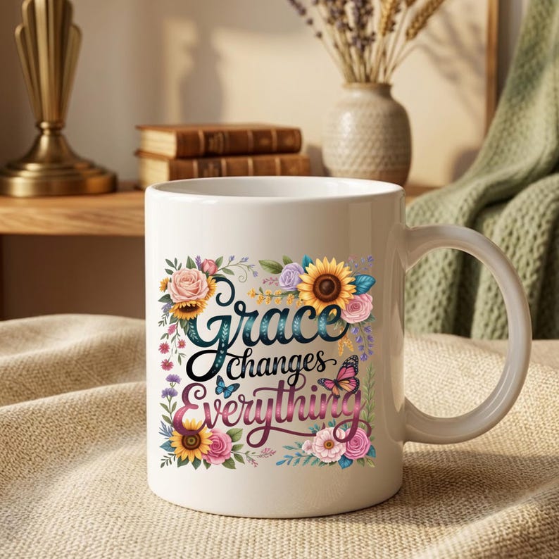 May include: A white ceramic mug featuring a colorful floral design with sunflowers, roses, and butterflies surrounding the text "Grace changes Everything" in a dark blue and pink script font, centered on the front of the cylindrical vessel.