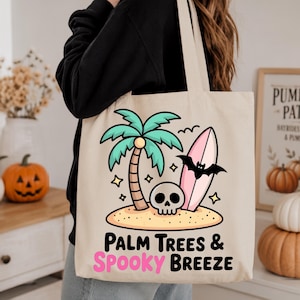 May include: A beige canvas tote bag featuring a graphic of a tropical island with a palm tree, a skull, and a pink surfboard with a black bat. Below the illustration, the text reads "PALM TREES & SPOOKY BREEZE" in black and pink lettering.