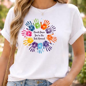 May include: White t-shirt featuring a colorful handprint design arranged in a circle. The design includes handprints in various colors, such as pink, green, orange, and purple, with the text "Hands Down You're the Best Around".