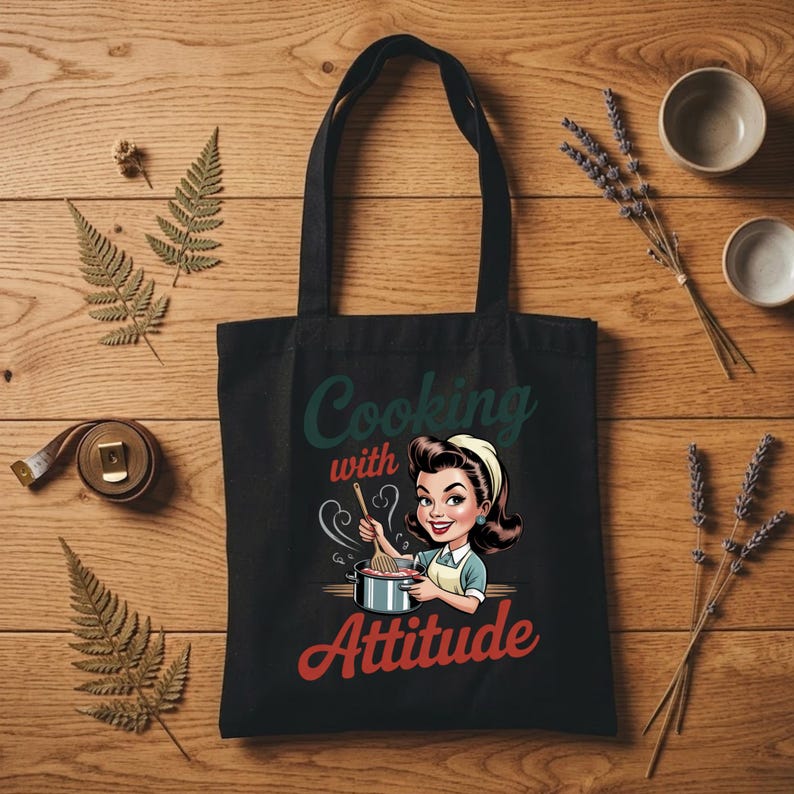 May include: Black tote bag with the text "Cooking with Attitude" and a retro cartoon image of a woman cooking. The bag has two black handles and is made of a sturdy material. The background is a wooden surface with decorative elements.