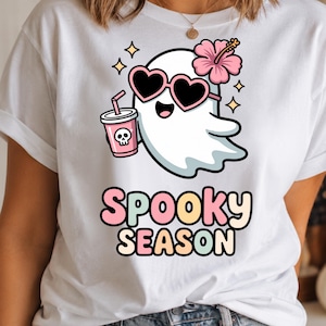 May include: A white t-shirt featuring a graphic of a cartoon ghost wearing pink heart-shaped sunglasses and a pink hibiscus flower. The ghost holds a pink cup with a skull icon. Below the ghost, the text reads Spooky Season in pastel colors.