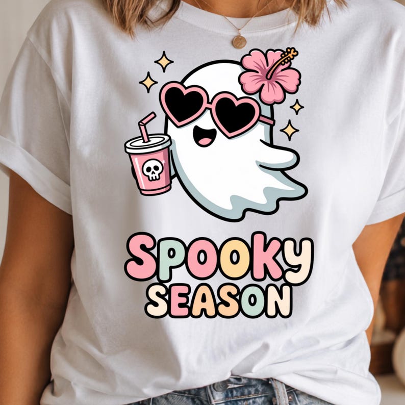 May include: A white t-shirt featuring a graphic of a cartoon ghost wearing pink heart-shaped sunglasses and a pink hibiscus flower. The ghost holds a pink cup with a skull icon. Below the ghost, the text reads Spooky Season in pastel colors.