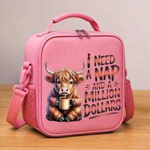 May include: A pink lunch bag with a top handle and a shoulder strap. The front features a graphic of a fluffy brown highland cow holding a mug, with the text "I NEED A NAP AND A MILLION DOLLARS" printed in a dark, stylized font next to the cow.