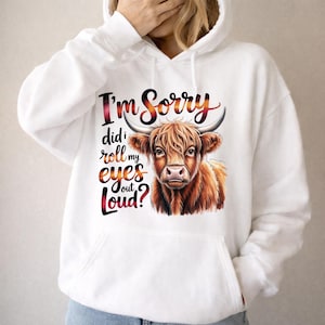 May include: A white hooded sweatshirt featuring a graphic of a brown Highland cow. The text printed in a red and orange gradient reads, I'm Sorry did I roll my eyes out Loud? The hoodie includes a front kangaroo pocket and long sleeves.