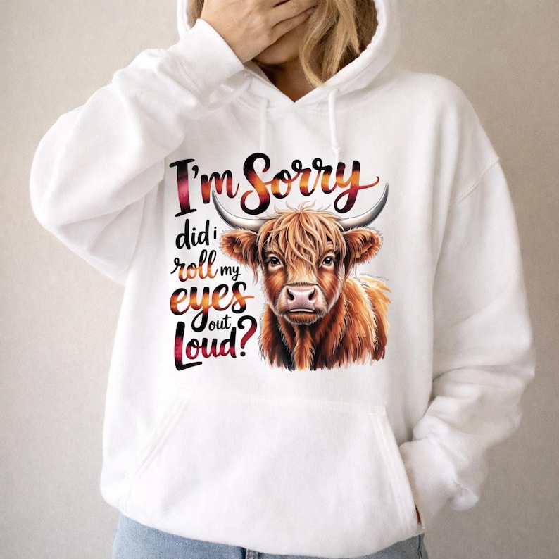 May include: A white hooded sweatshirt featuring a graphic of a brown Highland cow. The text printed in a red and orange gradient reads, I'm Sorry did I roll my eyes out Loud? The hoodie includes a front kangaroo pocket and long sleeves.