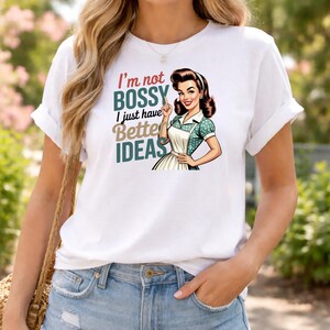 May include: White t-shirt with the text "I'm not bossy, I just have better ideas" and a retro illustration of a woman in an apron. The woman is pointing upwards. The shirt has rolled-up sleeves.