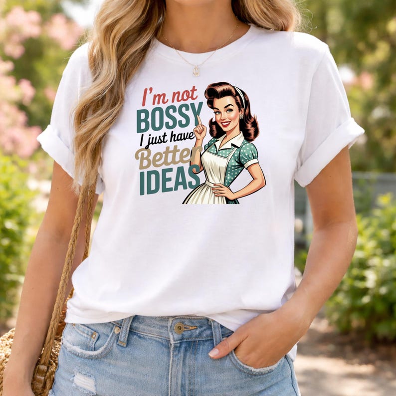 May include: White t-shirt with the text "I'm not bossy, I just have better ideas" and a retro illustration of a woman in an apron. The woman is pointing upwards. The shirt has rolled-up sleeves.