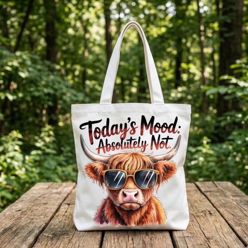 May include: A white canvas tote bag featuring a graphic illustration of a shaggy brown Highland cow wearing yellow-rimmed sunglasses. Above the cow, the text reads Today's Mood: Absolutely Not. The bag has two shoulder straps and a rectangular shape.