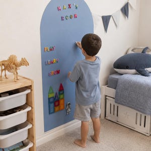 以下が含まれることがあります： 子供が、カラフルな文字で「MAX'S ROOM」と「PLAY, LEARN, GROW」と書かれた青い壁の前に立っています。マグネット式のブロックと宇宙飛行士のフィギュアが壁に取り付けられています。木製のおもちゃの恐竜と収納ユニットが背景にあります。
