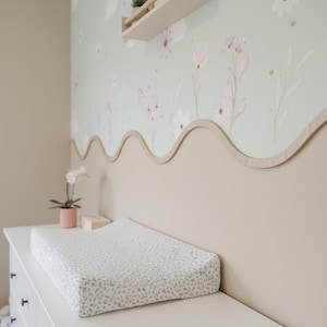 May include: A white changing pad with a gray leaf pattern rests on a white dresser. The wall features a floral wallpaper with pink flowers and a light green background. A wooden trim separates the wallpaper from the beige wall.