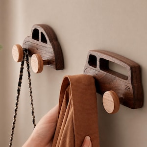 May include: Two dark brown wooden wall hooks shaped like vehicles, each featuring a lighter wood circular knob as a wheel. One hook holds a thin black chain, while the other supports a folded brown fabric item, showcasing the functional design.