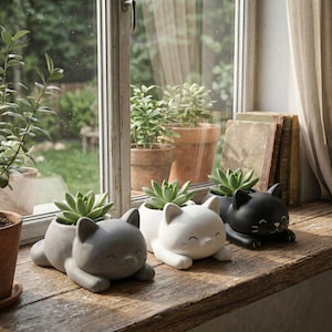 Peut inclure: Trois pots de fleurs en forme de chat, gris, blanc et noir, chacun contenant une plante succulente verte. Les pots sont sur un rebord de fenêtre en bois, avec une fenêtre et des plantes en arrière-plan.