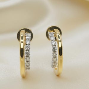 May include: A pair of gold-coloured hoop earrings. Each earring has a double hoop design, with one hoop set with small, clear gemstones. The earrings are displayed against a soft, off-white background.
