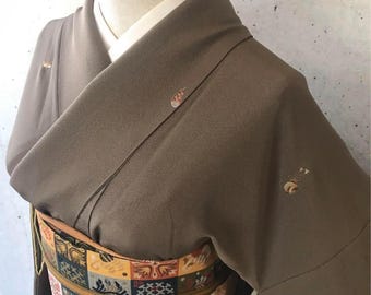 May include: A brown kimono with a patterned obi belt. The kimono has embroidered details on the sleeves and shoulders. The obi belt features a colorful geometric design with squares of various colors and patterns.