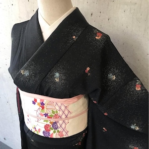 May include: Black kimono with floral accents and a patterned obi. The kimono features small, colorful floral designs scattered across the fabric. The obi is cream-colored with a pink border and a repeating floral pattern.