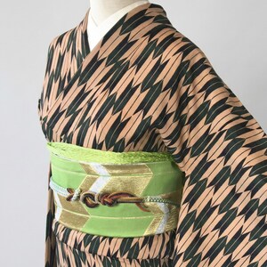 May include: A traditional Japanese kimono with a tan and black geometric pattern. The kimono is cinched at the waist with a bright green obi belt with gold and white accents. The obi is tied with a decorative knot.