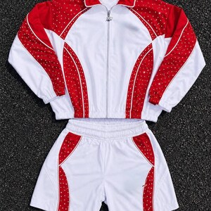 May include: A two-piece set consisting of a white and red zip-up track jacket and matching shorts. The jacket features a collar, long sleeves, and red panels adorned with small white dots. The shorts have an elastic waistband and matching red panels.