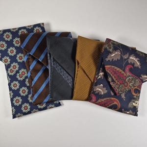 May include: A collection of five folded neckties featuring diverse patterns and colors. The set includes a navy blue floral print, a brown tie with blue diagonal stripes, a solid dark gray, a textured mustard yellow, and a navy paisley print.