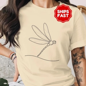Puede incluir: Camiseta color crema con un dibujo lineal minimalista de una libélula en vuelo. La libélula tiene alas delicadas y un cuerpo delgado. Una insignia roja con el texto "SHIPS FAST" es visible.