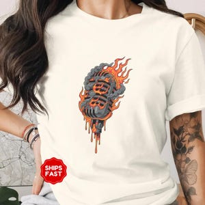 May include: Cream-colored t-shirt featuring a graphic of a vintage microphone engulfed in flames and smoke. The microphone is black and gray with orange flames and dripping orange accents. The shirt has short sleeves.