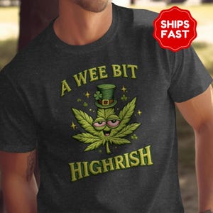 Puede incluir: Camiseta gris oscuro con un personaje de hoja de cannabis verde con sombrero de copa y el texto "A Wee Bit Highrish" en amarillo. El diseño incluye tréboles y estrellas.
