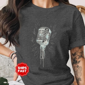 May include: Dark gray t-shirt featuring a vintage microphone illustration in shades of silver and light blue, surrounded by delicate leaf outlines. The design is centered on the shirt. The shirt is a crew neck.