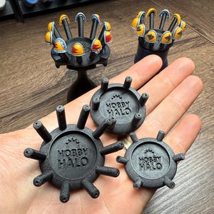 May include: Black 3D-printed Hobby Halo paint handle bases in various sizes. The bases have multiple posts for holding miniature figures. Two black stands hold painted miniature figures with yellow and orange helmets. The text "HOBBY HALO" is printed on the bases.