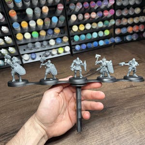 May include: A black miniature figure holder with five unpainted figurines of armoured warriors, each holding a weapon or shield. The background shows a collection of paint bottles.